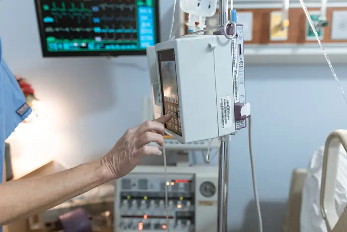 A gloved hand setting an infusion pump beside a monitor showing waveform traces